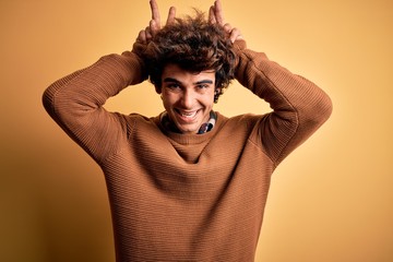 Young handsome man wearing casual shirt and sweater over isolated yellow background Posing funny and crazy with fingers on head as bunny ears, smiling cheerful