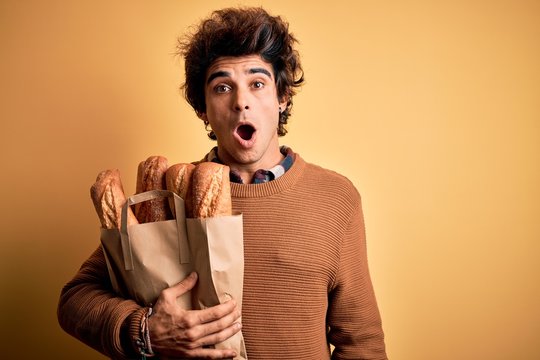 Young handsome man holding paper bag with bread standing over isolated yellow background scared in shock with a surprise face, afraid and excited with fear expression