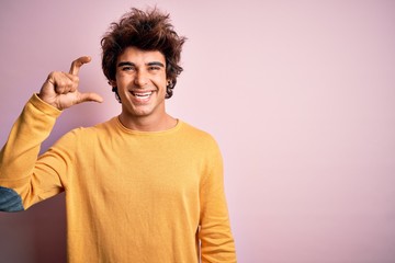 Young handsome man wearing yellow casual t-shirt standing over isolated pink background smiling and confident gesturing with hand doing small size sign with fingers looking and the camera. Measure