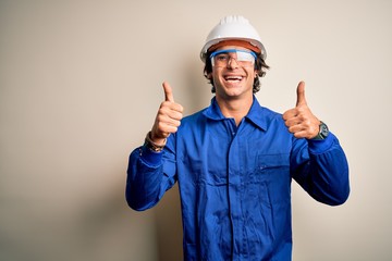 Young constructor man wearing uniform and security helmet over isolated white background success sign doing positive gesture with hand, thumbs up smiling and happy. Cheerful expression 