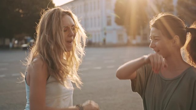 Outdoor portrait of two hipster friends with skateboard high five in sunshine city. Two fashion hipster women walking with coffee in hands at hot summer day and skateboarding lifestyle.