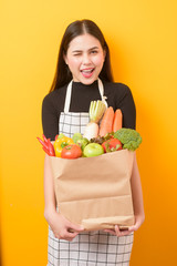  Beautiful young woman is holding vegetables in grocery bag in studio yellow  background