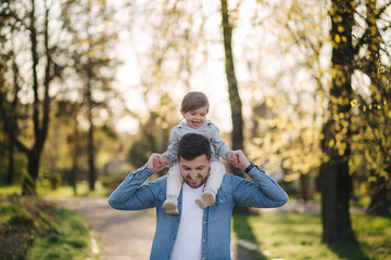 Adorable little daughter sitting on dad's neck and laughing. Young father walk with his cute daughter in the park