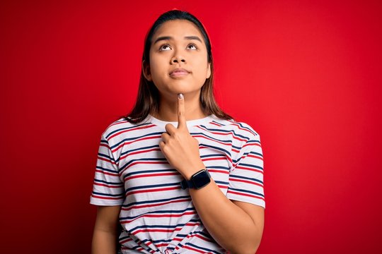 Young beautiful asian girl wearing casual striped t-shirt over isolated red background Thinking concentrated about doubt with finger on chin and looking up wondering
