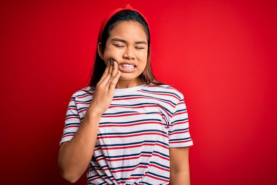 Young beautiful asian girl wearing casual striped t-shirt over isolated red background touching mouth with hand with painful expression because of toothache or dental illness on teeth. Dentist