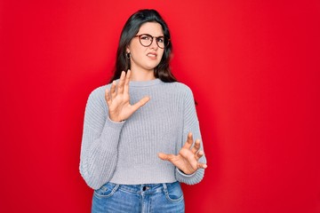 Young beautiful brunette woman wearing casual sweater over red background disgusted expression,...