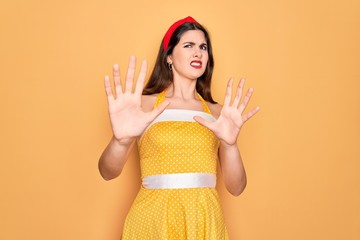 Young beautiful pin up woman wearing 50s fashion vintage dress over yellow background afraid and terrified with fear expression stop gesture with hands, shouting in shock. Panic concept.