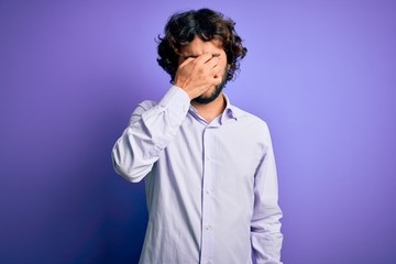 Young handsome business man with beard wearing shirt standing over purple background tired rubbing nose and eyes feeling fatigue and headache. Stress and frustration concept.