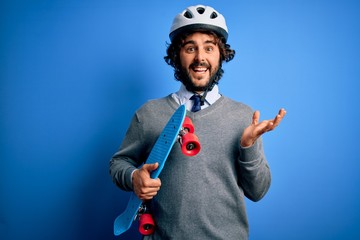 Handsome skater man with beard wearing security helmet holding skate over blue background very happy and excited, winner expression celebrating victory screaming with big smile and raised hands