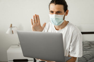 Man in medical mask is working from home with a laptop and speaking to his friends or business partners via webcam because of coronavirus.