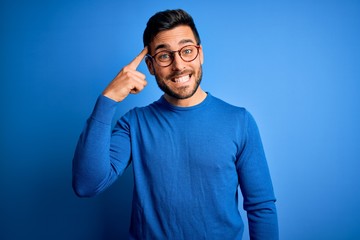 Young handsome man with beard wearing casual sweater and glasses over blue background Smiling...