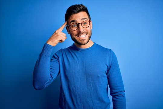 Young handsome man with beard wearing casual sweater and glasses over blue background Smiling pointing to head with one finger, great idea or thought, good memory