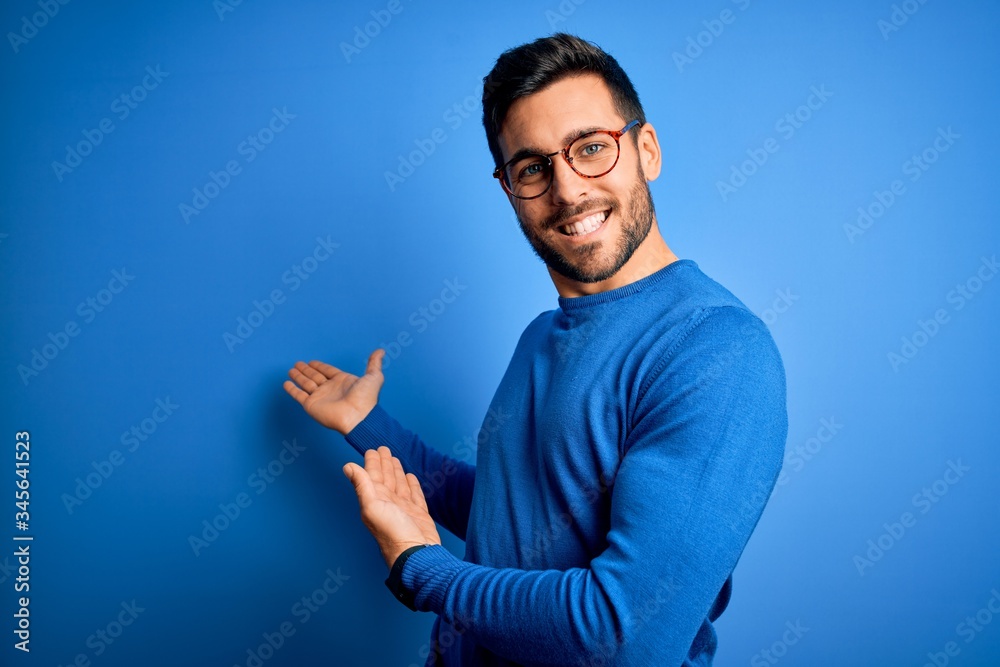 Poster young handsome man with beard wearing casual sweater and glasses over blue background inviting to en