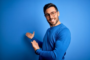 Young handsome man with beard wearing casual sweater and glasses over blue background Inviting to enter smiling natural with open hand
