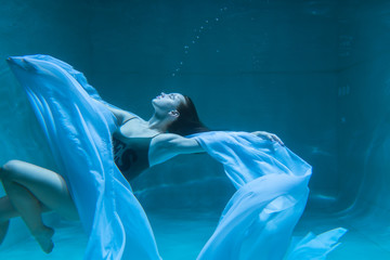 Beautiful girl swims underwater with long hair. Blue or gold background like gold. The atmosphere of a fairy tale or magic. Diving under the water with a shiny cloth