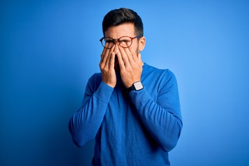 Young handsome man with beard wearing casual sweater and glasses over blue background rubbing eyes for fatigue and headache, sleepy and tired expression. Vision problem