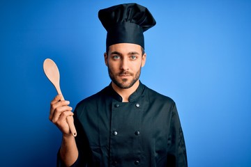 Naklejka premium Young cooker man with beard wearing uniform holding wooden spoon over blue background with a confident expression on smart face thinking serious