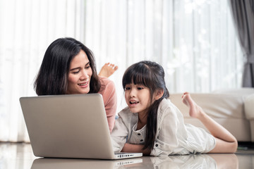 Fototapeta premium Homeschool Asian little young girl feel relax, learning internet online class by using computer with mother help, teach laying on floor. Daughter smile and happy to study at home together with mom.