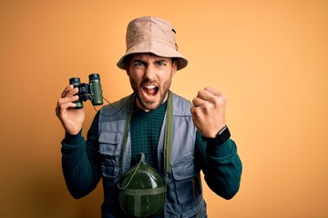 Young handsome tourist man with beard on vacation wearing explorer hat using binoculars annoyed and frustrated shouting with anger, crazy and yelling with raised hand, anger concept