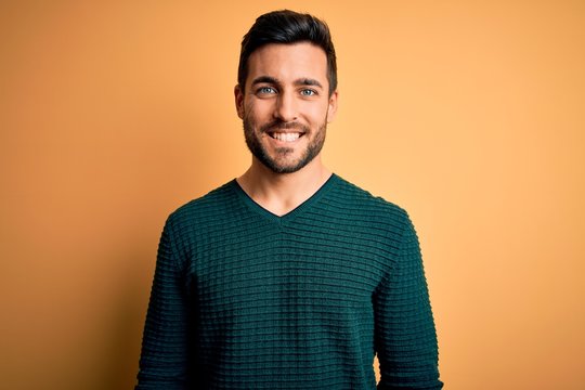 Young handsome man with beard wearing casual sweater standing over yellow background with a happy and cool smile on face. Lucky person.