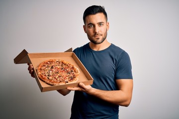 Young handsome man holding delivery box with delicious Italian pizza over white background with a confident expression on smart face thinking serious