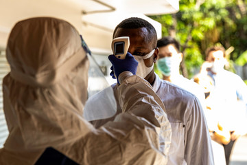 Security guard in PPE suit uses infrared thermometer measuring temperature with international staff...