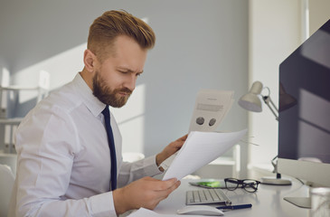 Serious busy businessman reads working documents sitting at a table with a computer in the office.