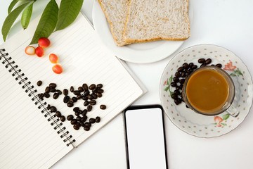 Top view of coffee beans and carissa carandas on notebook,leaves,two pieces of bread,mobile phone,and cup of coffee with saucer,isolated on background.