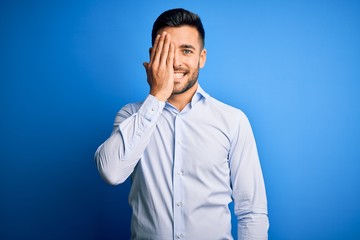 Young handsome man wearing elegant shirt standing over isolated blue background covering one eye with hand, confident smile on face and surprise emotion.