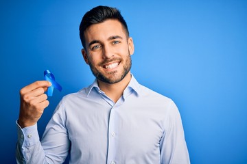 Young handsome man holding blue ribbon as prostate campaing support over blue background with a...