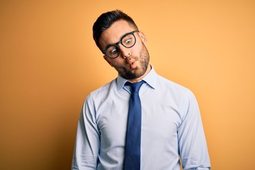 Young handsome businessman wearing tie and glasses standing over yellow background making fish face with lips, crazy and comical gesture. Funny expression.
