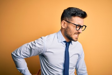 Young handsome businessman wearing tie and glasses standing over yellow background Suffering of backache, touching back with hand, muscular pain