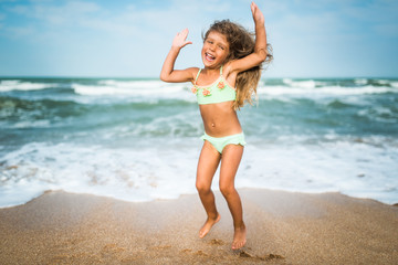 Fototapeta premium Cheerful little caucasian girl jumping and having fun on the sea waves on the sandy coast on a sunny hot summer day. Concept of vacation with children as a family
