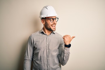 Young architect man wearing builder safety helmet over isolated background smiling with happy face looking and pointing to the side with thumb up.