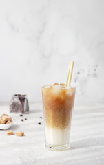 Cold coffee with milk and ice cubes in a tall glass with bamboo straw, light grey background. Selective focus. 