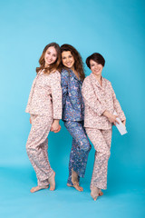 Portrait of three beautiful young girls wearing colorful pyjamas having fun during sleepover isolated over blue background. Pajama party and hen-party concept