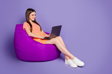 Full length profile photo of cheerful lady sitting comfy beanbag browsing notebook freelance work wear orange striped t-shirt jeans mini skirt isolated pastel purple color background