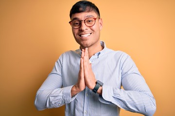 Young handsome hispanic business man wearing nerd glasses over yellow background praying with hands...