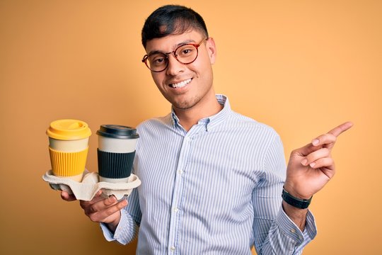 Young scholarship holder business man holding take away coffee over isolated yellow background very happy pointing with hand and finger to the side