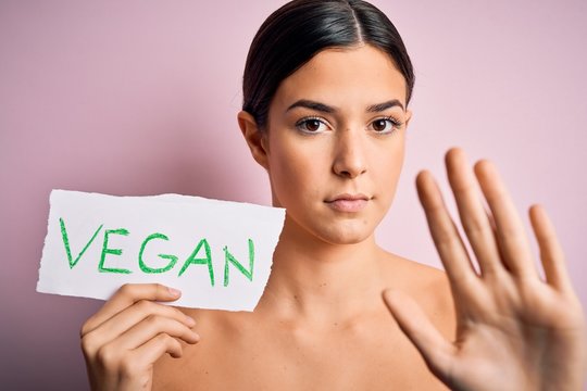 Young beautiful girl holding paper with vegan message over isolated pink background with open hand doing stop sign with serious and confident expression, defense gesture