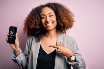 Young african american woman with afro hair holding cracked and broken smartphone screen very happy pointing with hand and finger