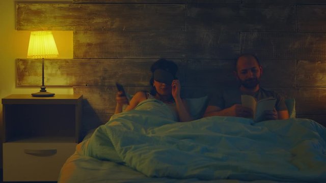 Wife turning the main lights in the room using a phone app while her husband is reading a book.