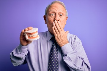 Grey haired senior man holding orthodontic prosthesis denture over purple background cover mouth with hand shocked with shame for mistake, expression of fear, scared in silence, secret concept - obrazy, fototapety, plakaty