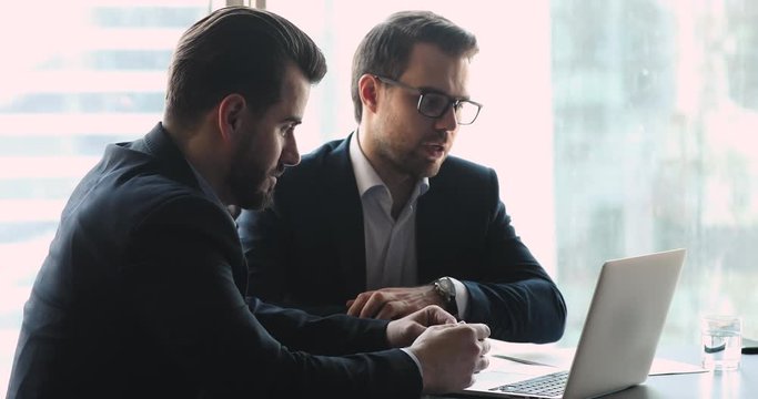 Two focused businessmen in formal suits sitting at table, watching discussing business ideas presentation on laptop in office. Successful financial advisor giving professional consultation to client.