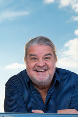 Portrait of a latin man when he is standing outside of a window. He is looking down at the camera while leaning the window pane. The sky serves as background. He is smiling.