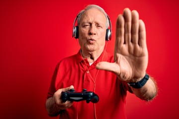 Senior grey haired gamer man playing video games using gamepad joystick over red background with open hand doing stop sign with serious and confident expression, defense gesture © Krakenimages.com