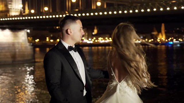 Young Wedding Couple, Bride And Groom In White Dress, Dancing, Holding Hands, Hugging, Against The Background Of Night Old Town, River And Big Bridge. Bokeh Lights. Light. Evening. Slow Motion.