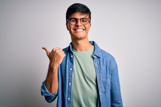 Young handsome man wearing casual shirt and glasses over isolated white background smiling with happy face looking and pointing to the side with thumb up.