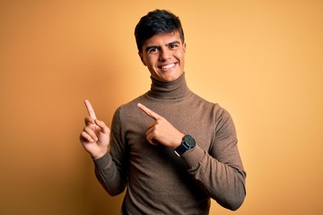 Young handsome man wearing casual turtleneck sweater over isolated yellow background smiling and looking at the camera pointing with two hands and fingers to the side.