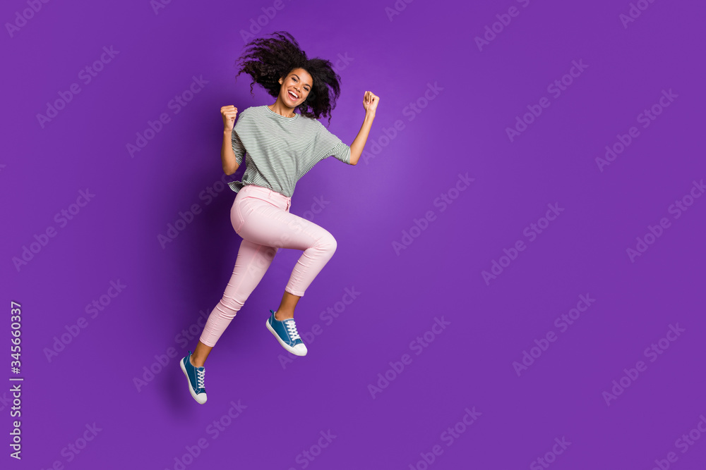 Poster Full length profile side photo of cheerful funky afro american girl jump enjoy discounts win lottery raise fists scream yes wear striped shirt sneakers isolated violet color background